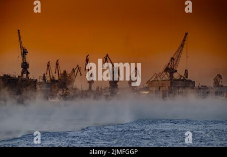 Der Bau von nuklearen Eisbrechern bei magischem Sonnenuntergang, Kräne der baltischen Werft in einem frostigen Wintertag, Dampf über die Stockfoto