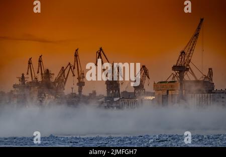 Der Bau von nuklearen Eisbrechern bei magischem Sonnenuntergang, Kräne der baltischen Werft in einem frostigen Wintertag, Dampf über die Stockfoto