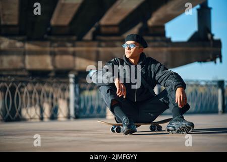 Skater männlich entspannen sitzen auf Skateboard auf Straße städtischen Hintergrund Stockfoto