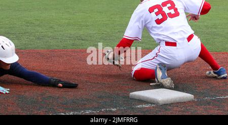 Der Kopf des Baseballspielers rutscht zuerst mit Gleithandschuhen in den dritten Platz, während der dritte Baseman versucht, den Tag zu verwenden. Stockfoto