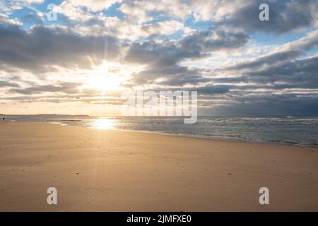Erstaunlicher farbenprächtiger Sonnenuntergang an einem Sandstrand am Meer unter einem Himmel, der mit Wolken und einer goldenen Sonne bemalt ist. Malerische Naturkulisse Stockfoto
