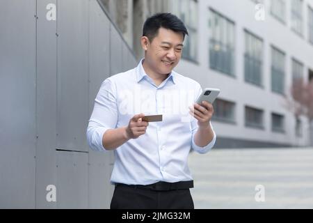 Asiatischer Geschäftsmann lächelt und macht gerne Online-Shopping vor dem Bürogebäude, Mann benutzt Mobiltelefon und hält Bankkreditkarte Stockfoto