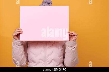 Frau in rosa Winterjacke und Hut hält rosafarbenes Blatt Papier auf gelbem Hintergrund. Saisonaler Verkauf Stockfoto