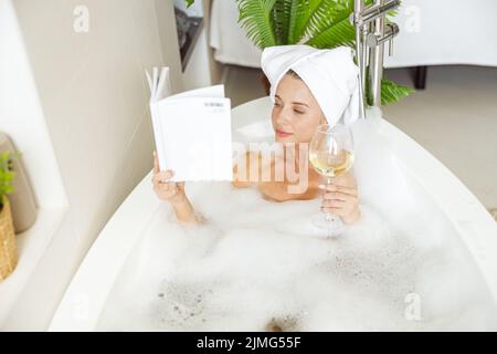 Schöne glückliche junge Frau, die mit Schaum im Bad liegt, Buch liest und Wein trinkt. Stockfoto