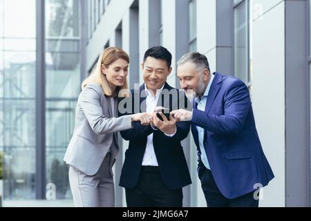 Vielfältiges Business-Team, Männer und Frauen vor dem Büro lächelnd und glücklich, schauend lustige Inhalte am Telefon Stockfoto