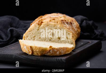 Gebackenes, ovales Vollbrot aus weißem Weizenmehl auf einem schwarzen Tisch, frisches Gebäck Stockfoto