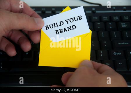 Hand halten mit Notizblock in gelbem Umschlag mit Text - bauen Sie Ihre Marke. Mit Hintergrund des Keyboard-Monitors. Branding-Konzept. Stockfoto