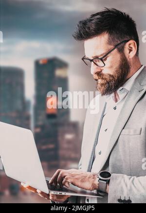 Erfolgreicher Geschäftsmann mit Laptop-Bürogebäuden im Hintergrund Stockfoto