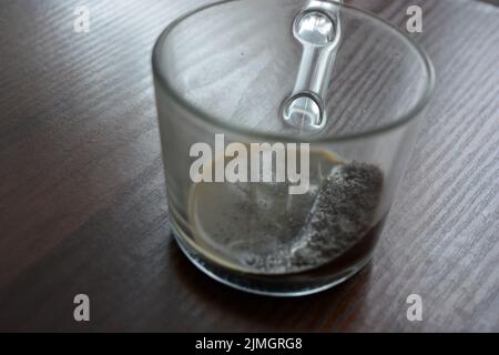 Auf einem dunkelgelben Holztisch befindet sich eine transparente, angenehme Glastasse mit natürlichem Kaffee und Milch. Stockfoto