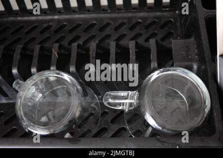 Zwei 2 gewaschene, klare Gläser-Tassen mit Besteck sind wunderschön auf einem schwarzen Kunststoff-Geschirrkorb angeordnet. Stockfoto