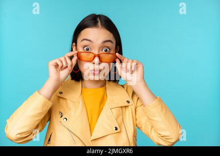 Nahaufnahme Porträt der asiatischen Mädchen in Sonnenbrille, schauen glücklich und überrascht, Aufregung im Gesicht, über blauem Hintergrund stehen Stockfoto