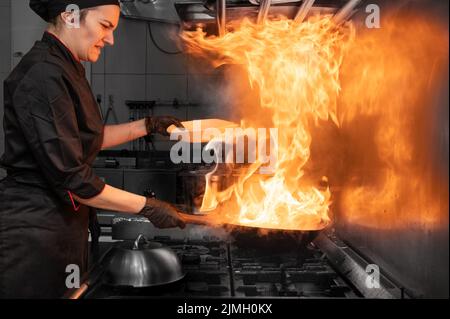 Frau Chefkoch Kochen Wok in der Küche. Kochen flammender Wok mit Gemüse in der kommerziellen Küche. Stockfoto