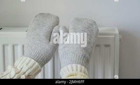 Nahaufnahme einer Frau in weißem Pullover und grauen Fäustlingen wärmt ihre Hände über einen Heizkörper an einer weißen Wand. Makrofoto von weiblichen Händen, Kopierraum. Gas Stockfoto