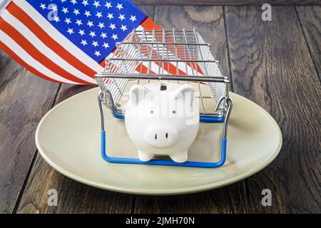 Sparschwein auf Teller unter dem Warenkorb und USA Flagge. Konzept der Handelsinflation Stockfoto