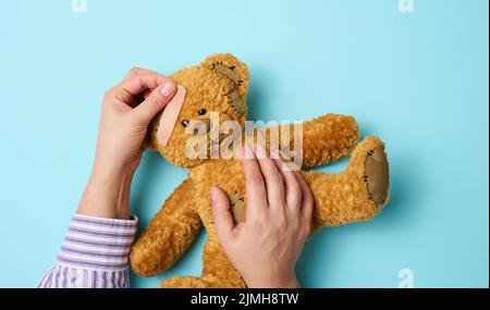 Weibliche Hand hält einen braunen Teddybär und klebt einen medizinischen Klebstoff-Gips auf blauem Hintergrund, Tram-Behandlung Stockfoto