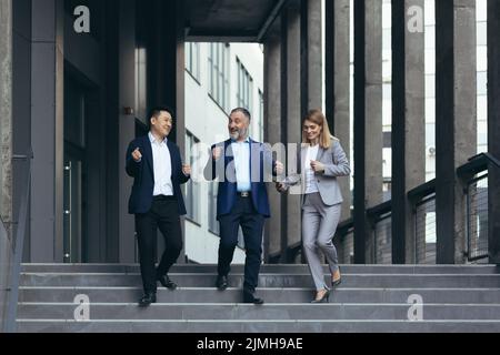 Ein vielfältiges Geschäftsteam, Männer und Frauen, die vor dem Bürogebäude zusammenlaufen, zufrieden mit der Siegerfeier, Geschäftsleute, die auf Treppen tanzen und glücklich lächeln Stockfoto