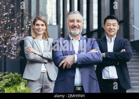 Vielfältiges Dream Team, leitender grauhaariger Geschäftsmann-Chef mit Kollegen vor dem Büro, die lächeln und mit gekreuzten Armen auf die Kamera blicken Stockfoto