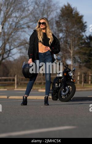 Ein schönes blondes Model genießt das Wetter im Freien, während sie mit ihrem Motorrad posiert Stockfoto