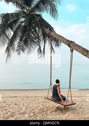 Frau, die auf einer Schaukel auf einer Palme sitzt und auf den ruhigen blauen Ozean am paradiesischen Sandstrand blickt Stockfoto