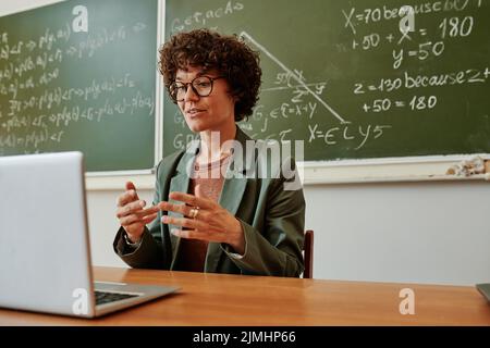 Selbstbewusster junger Lehrer, der neue Themen erklärt oder Fragen des Online-Publikums beantwortet, während er am Arbeitsplatz vor dem Laptop sitzt Stockfoto