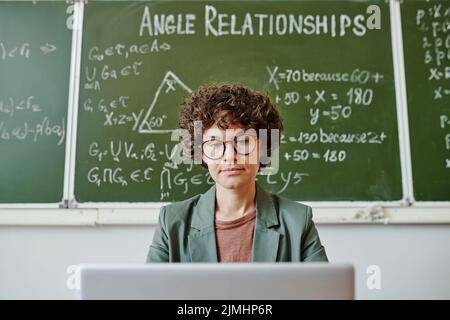 Junge ernsthafte Lehrerin in intelligenter Casualwear, die beim Sitzen an der Tafel mit Formeln und Gleichungen beim Unterricht auf den Laptop-Bildschirm schaut Stockfoto