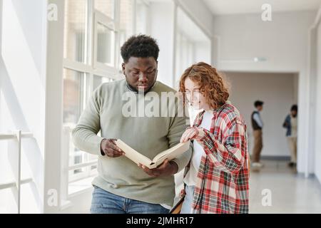 Zwei interracial Teenager Studenten in Casualwear lesen Buch aus der Bibliothek, während sie entlang College-Korridor vor dem Unterricht Stockfoto