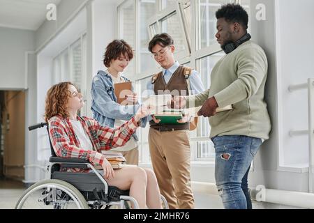 Nettes Teenager-Mädchen mit Behinderung nimmt Copybook von afroamerikanischen Kerl vor ihr stehen, während ihre Klassenkameraden mit Diskussion Stockfoto