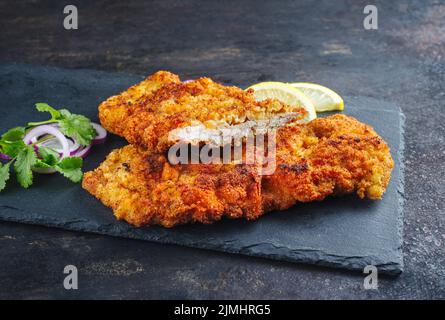 Traditionelles frittiertes Schnitzel mit Zitronenscheiben und Zwiebelringen, das als Nahaufnahme auf einem schwarzen Tablett serviert wurde Stockfoto