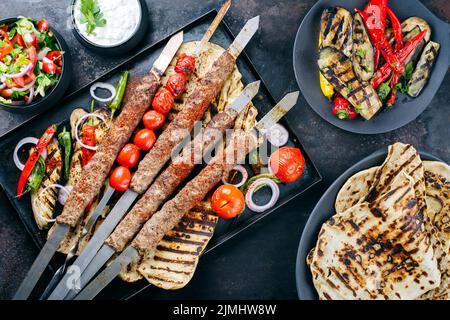 Traditioneller türkischer Adana-Kebap auf Schaschlik-Spieß mit Grillgemüse und Fladenbrot, serviert als Draufsicht auf einem rustikalen Metalltablett Stockfoto