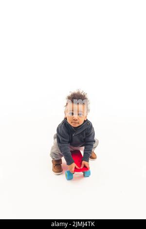 Ein kleiner fünf Jahre alter afroamerikanischer Kerl, der auf einem Skateboard sitzt und in die Kamera schaut - weißer Hintergrund. Hochwertige Fotos Stockfoto