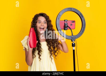 Teen girl, das vor der Kamera für Vlog auf gelbem Hintergrund isoliert spricht. Teenager arbeiten als Blogger halten Haarspülung oder Shampoo-Flasche Stockfoto