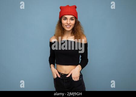 Foto von ziemlich positiv lächelnd junge Brunet lockig Frau isoliert über blauen Hintergrund Wand trägt schwarzen Crop Top und stilvoll Stockfoto