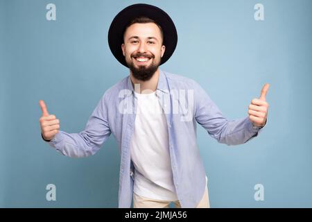 Foto von einem positiven fröhlichen lächelnden, gut aussehenden jungen Bärtigen mit Brunet, der ein legeres blaues Hemd und ein weißes T-Shirt und ein weißes Styli trägt Stockfoto