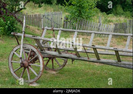 Alte Holzwagen in Serbien ruht auf dem Rasen. Sie können die alte handgemachte Bremse sehen. Ein Holzzaun mit grünen Büschen ist im Hintergrund. Stockfoto