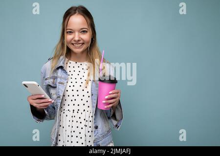 Foto Nahaufnahme der wunderschönen hübschen Frau in jean Jacke lächeln und halten Smartphone beim Trinken zum Mitnehmen Kaffee isoliert Ove Stockfoto