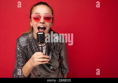 Schuss von schönen emotionalen positiv glücklich Brünette weibliche Teenager trägt stilvolle Glanz Jacke und rote Brille isoliert stehend Stockfoto