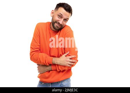 Schüchterner bärtiger Mann in einem stilvollen orangefarbenen Langarm schließt schüchtern seine Hände vor dem Hintergrund einer weißen Wand Stockfoto