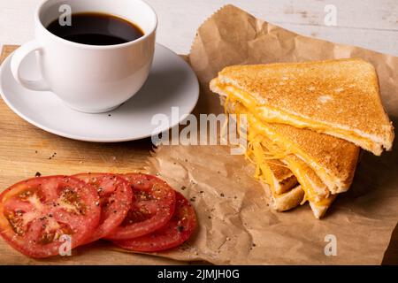 Blick aus der Perspektive auf frischen schwarzen Kaffee mit Tomatenscheiben durch Käse-Sandwich auf Wachspapier Stockfoto