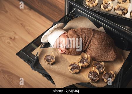 Das neugeborene Baby mit Kochmütze liegt mit Muffins auf dem Backblech Stockfoto