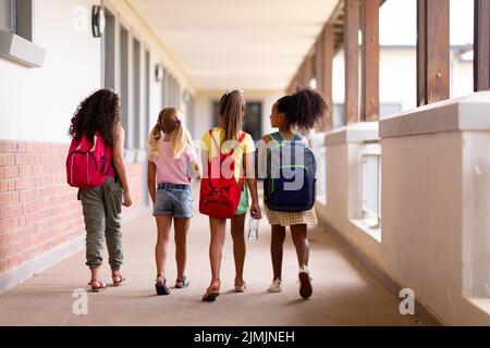 Lange Rückansicht von multirassischen Grundschulmädchen mit Rucksäcken, die im Flur laufen Stockfoto