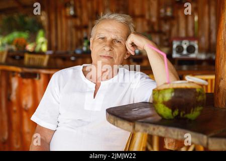 Ein glücklicher älterer Mann trinkt ein Kokoswasser in der Strandbar Stockfoto