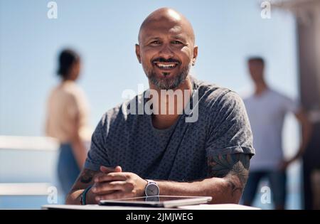 Ich gehe gerne ganz allein hierher. Porträt eines fröhlichen Mannes mittleren Alters, der an einem Tisch vor einer Strandpromenade sitzt. Stockfoto