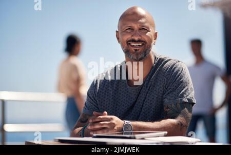 So machen Sie eine Pause vom Büro. Porträt eines fröhlichen Mannes mittleren Alters, der an einem Tisch vor einer Strandpromenade sitzt. Stockfoto