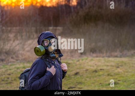 Ein Mädchen in einer schwarzen Gasmaske und dunkler Kleidung mit einer Kapuze bei Sonnenuntergang vor dem Hintergrund eines Frühlingswaldes. Stockfoto