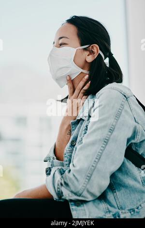 Eine Halsschmerzen ist ein häufiges Symptom von Covid-19. Eine junge Frau, die eine Maske trägt und in einer Arztpraxis unter Rachenschmerzen leidet. Stockfoto