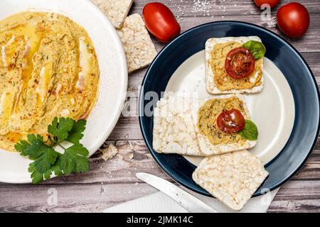 Quadratische Reiscracker mit Hummus und Kirschtomaten auf einem Teller. Tomaten, Cracker auf einem rustikalen Tisch und eine Platte mit Hummus. Natürlich und gesund f Stockfoto