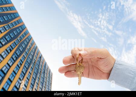 Wohnungsschlüssel in der Hand eines Mannes. Türschloss aus Messing. Modernes Gebäude am blauen Himmel. Ansicht von unten. Stockfoto