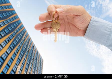 Wohnungsschlüssel in der Hand eines Mannes. Türschloss aus Messing. Modernes Gebäude am blauen Himmel. Ansicht von unten. Stockfoto