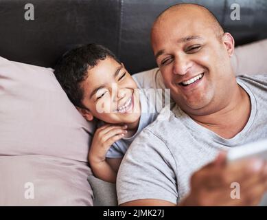 Sie sind immer lachend gelacht: Ein fröhlicher junger Mann und sein Sohn schauen sich gemeinsam Videos auf einem digitalen Tablet an, während sie zu Hause auf einem Bett rumhängen Stockfoto