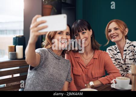 Alle lächeln. Eine Gruppe attraktiver Frauen mittleren Alters, die zusammen ein Selbstporträt machen, während sie tagsüber in einem Café sitzen. Stockfoto